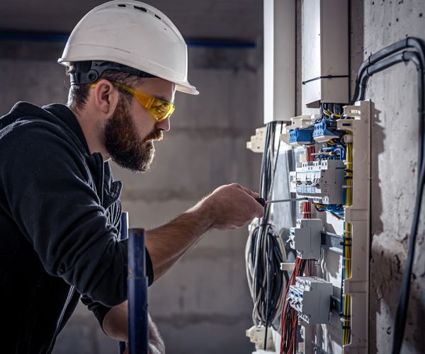 A male electrician works in a switchboard with an electrical connecting cable, connects the equipment with tools.